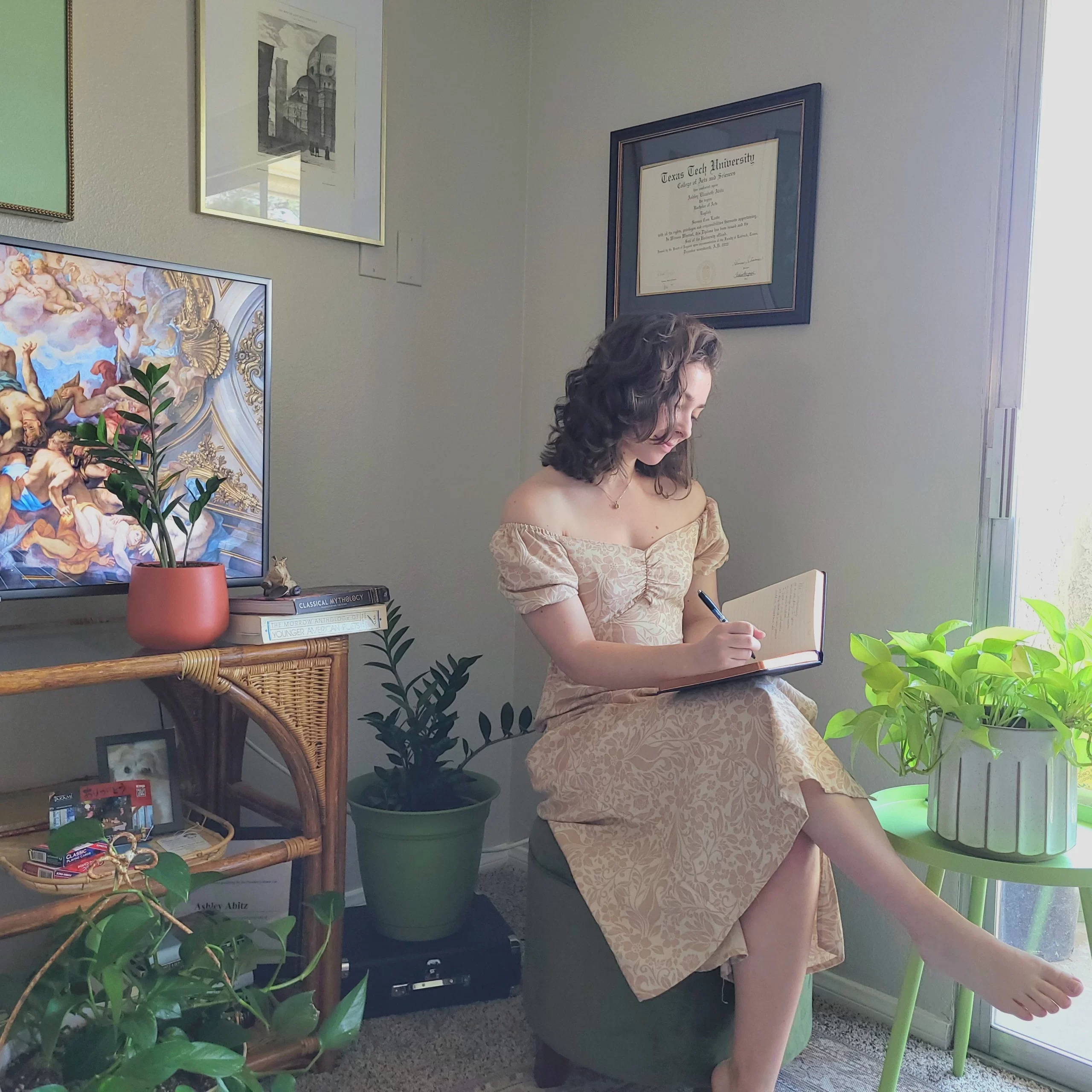 Young woman in an elegant off-the-shoulder dress sitting near a window, writing in a journal. She is surrounded by indoor plants and art.